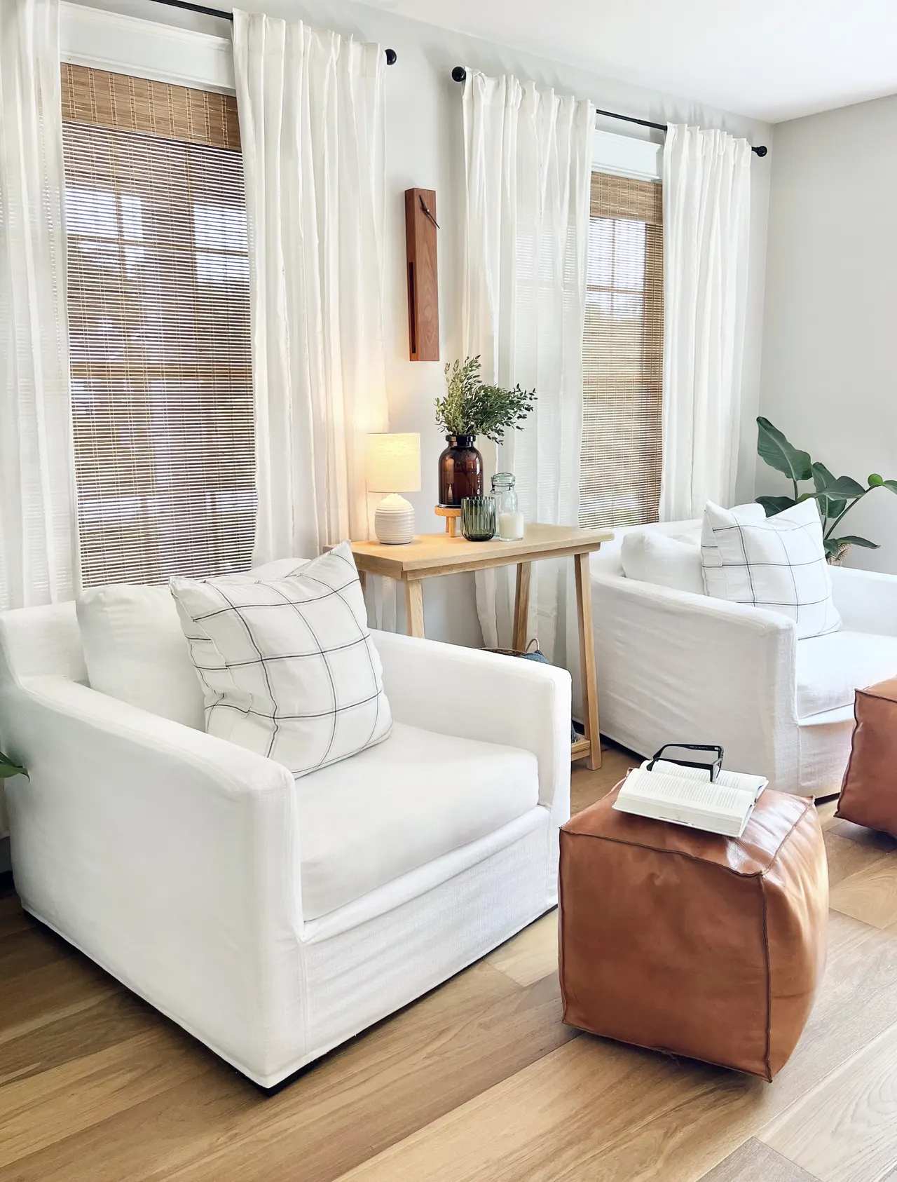 A sitting area with two white slipcovered chairs, windowpane pillows, leather poufs, and a wood side table by woven-shaded windows, designed by STELLAN